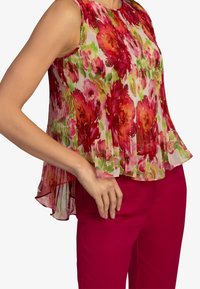 Blouse sans manches en tissu léger plissé. Présente des fleurs rouges et roses vives sur un fond clair, assortie à un pantalon rose.