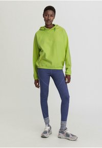 Limonengrüner Kapuzenpullover, blaue Leggings mit kontrastierenden Bündchen und graue Sportschuhe mit hellen Akzenten. Einfaches Design, bequemer Schnitt.