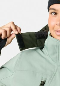 Zielona kurtka zewnętrzna z wysokim kołnierzem, wyposażona w czarny rzep. Gładka tekstura i dopasowany krój podkreślające funkcjonalne detale projektu.