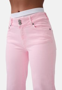 Jeans in denim rosa con vita alta, dotati di chiusura a pulsante, tasche frontali e dettagli di cucitura sottili. Visibile cinturino bianco con brand.