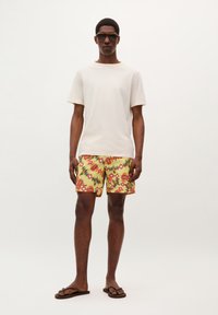 T-shirt écru à manches courtes, shorts à motifs vibrants en jaune avec des accents rouges et verts, tongs marron et lunettes de soleil portées par un mannequin.