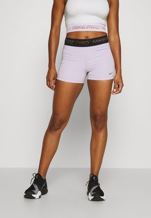 Junge Person trägt hellviolette Nike Pro Shorts, weißen Sport-BH und schwarze Sportschuhe, steht mit gespreizten Beinen vor einfarbigem Hintergrund.