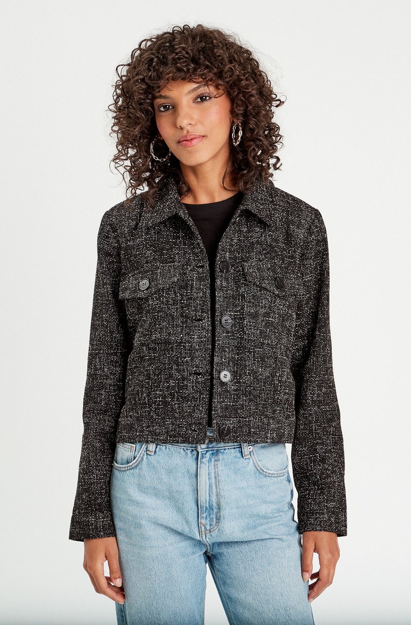 Giacca cropped in tweed bianco e nero con colletto, due tasche frontali e cinque bottoni, abbinata a jeans azzurri.