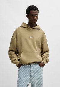Khaki hoodie van textiel met een structuur, ruime pasvorm en centraal logo-patch. Gecombineerd met licht blauwe geruite denim jeans.