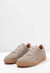 Beige mockasin-sneakers med perforerade detaljer, platt snörning och gummisula. Insidan har en kontrasterande brun foder.