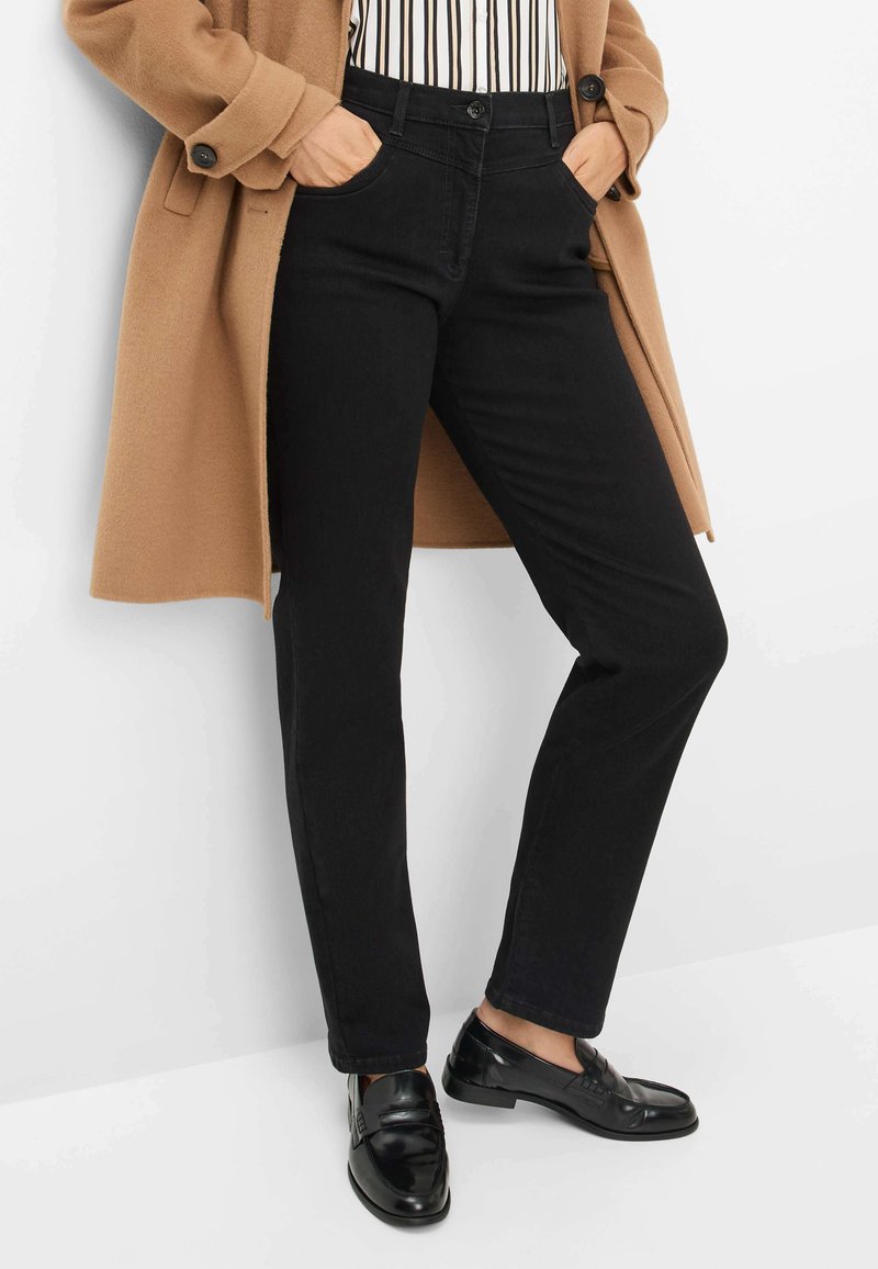 Jeans neri a vestibilità slim, cinque tasche, chiusura con bottone e leggero elastico, abbinati a un cappotto tan e mocassini neri lucidi.