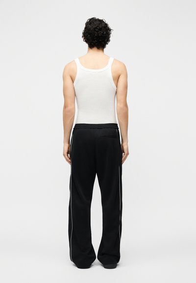 Άτομο με μπούκλες που φαίνεται από πίσω, φορώντας λευκό αμάνικο τοπ και μαύρο παντελόνι με φαρδιές λεκάνες και λευκές πλευρικές ρίγες, που στέκεται μπροστά σε απλό φόντο.