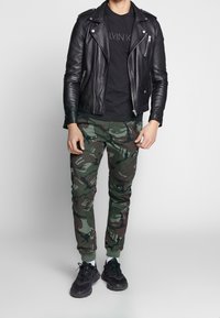 Svart läderbikerjacka över en svart t-shirt, ihopparat med gröna kamouflagebyxor som är avsmalnande och svarta sneakers med texturerade sulor.