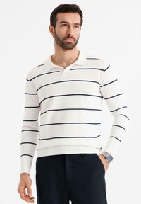 Muž v bílé polokošili s dlouhými rukávy a námořnickými horizontálními pruhy, tmavých kalhotách a stříbrných hodinkách, pózuje s jednou rukou v kapse.