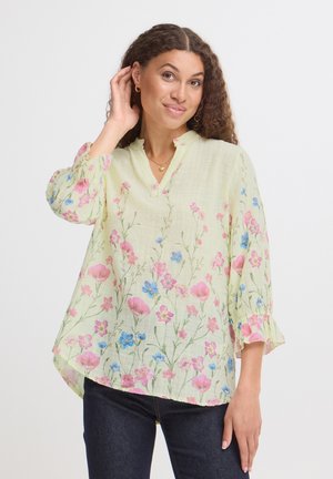 Kvinde med krøllet mørkt hår iført en lysegul bluse med lyserøde og blå blomsterprint og mørke jeans, der rører ved sit øre med den ene hånd.