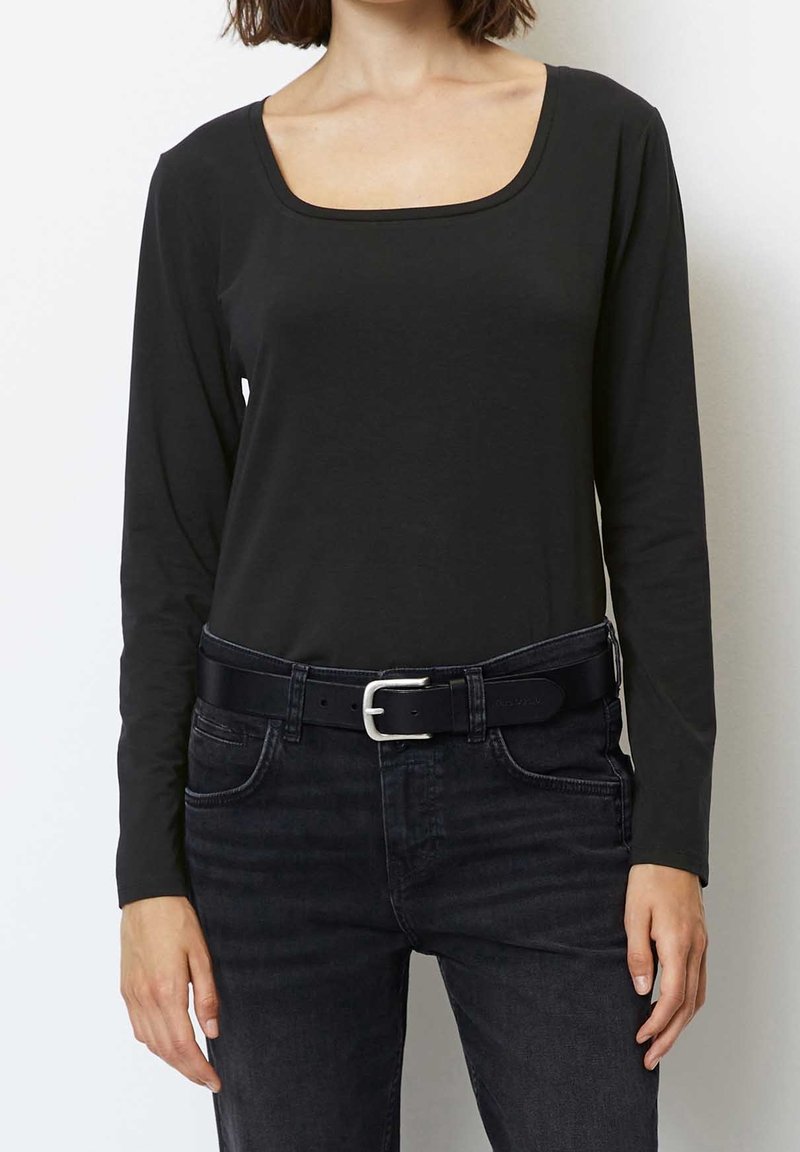 Kvinna som bär en svart långärmad topp med rund hals, svarta jeans och ett svart bälte med en silverspänne, stående mot en vit bakgrund.