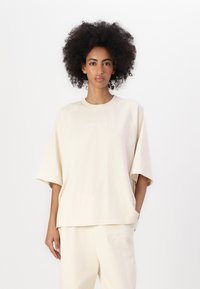Beige, oversized sweatshirt med korte ermer, rund halslinning og subtil brodering foran. Laget av myk bomull med en glatt tekstur.