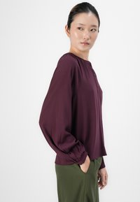 Bordová blůza s kulatým výstřihem a knoflíkovou légou. Má dlouhé, nabírané rukávy s elastickými manžetami. V kombinaci se zelenými kalhotami.