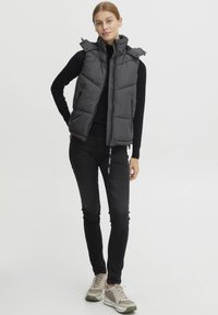 Veste grise rembourrée avec un col haut, fermeture éclair à l'avant et poches latérales, associée à un jean skinny noir et des baskets de couleur claire.