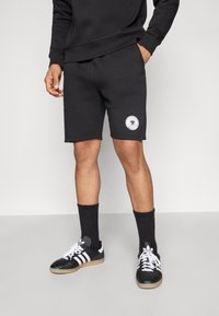 Musta urheilushortsit pyöristetyllä helmalla, joissa on valkoinen pyöreä logo. Päällä mustat sukat ja mustavalkoiset lenkkarit.