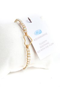 Goldfarbenes Drahtarmband mit kleinen Aquamarin-Perlen, herzförmigem Verschluss und einem angebrachten Etikett mit Text. Der Text lautet "Aquamarin", "Fluidität" und "Geh mit dem Fluss."