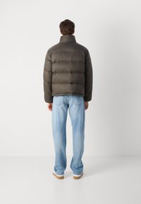 Doudoune vert olive foncé avec un col montant, design matelassé et poignets élastiques ; associée à un jean droit bleu clair et des chaussures beiges.