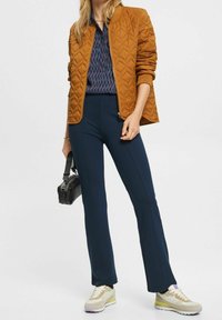 Giubbotto bomber marrone trapuntato, pantaloni fitted blu navy, camicia a fantasia e sneakers beige con accenti viola. Tiene una borsa nera.
