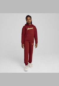 Sudadera burdeos con el logo dorado de "Puma", bolsillo canguro, combinada con pantalones a juego. Tejido suave, corte relajado, complementada con zapatillas blancas.