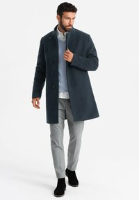 Cappotto di lana blu scuro con colletto alto, sette bottoni, abbinato a un maglione azzurro chiaro e pantaloni grigi, con stivaletti neri alla caviglia.