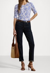 Kvinna klädd i en blå blommig topp, mörka uppvikta jeans med brunt bälte, bruna högklackade sandaletter, holding en väskan i två nyanser av brunt.