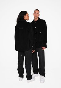 Zwarte corduroy jacks met drukknoopjes, gecombineerd met donkere denim broeken en witte sneakers. Textuur van de stof en details van de zakken zichtbaar.