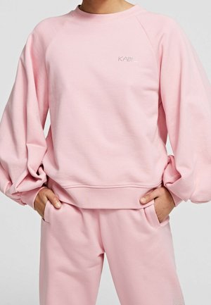 Personne portant un sweat-shirt ample rose clair et un pantalon assorti, les mains dans les poches, avec un petit détail de texte sur la poitrine.