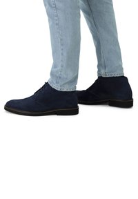 Bottines en cuir suede marine avec un bout arrondi et un design de lacets minimaliste, associées à un jean tapered bleu clair, avec une semelle noire lisse.