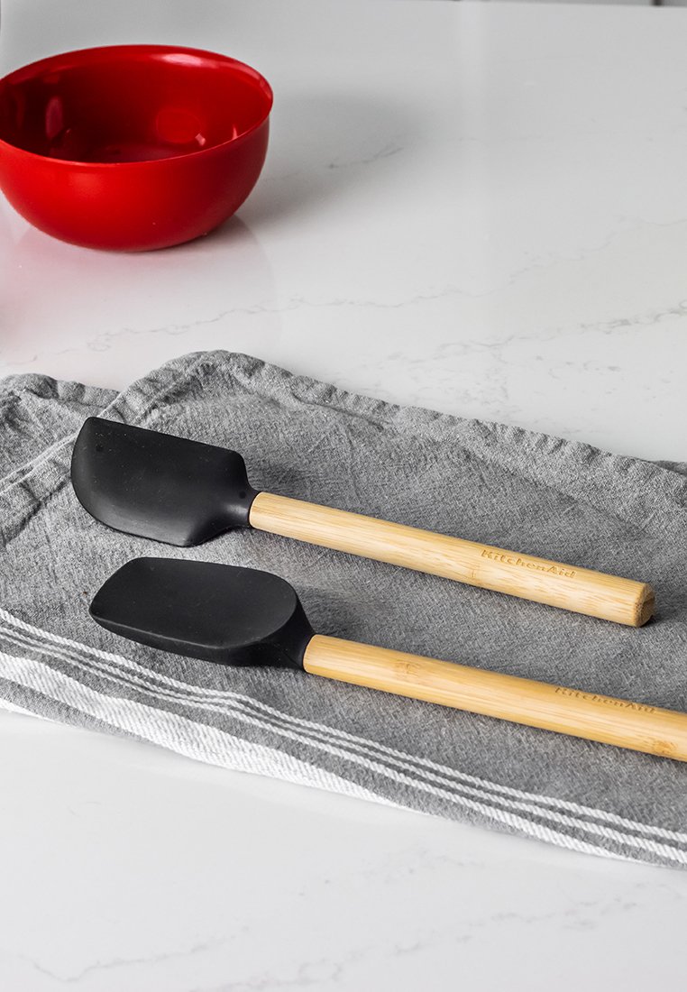 Twee zwarte siliconen spatels met houten handvatten op een grijze doek naast een rode kom op een wit aanrecht.