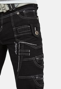 Pantaloni cargo neri con cuciture bianche, numerose tasche e hardware a pressione. Presentano una finitura testurizzata e un design aderente.