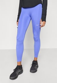 Μπλε αθλητικό κολάν από ελαστικό ύφασμα, με εφαρμοστή γραμμή και μικρό λευκό λογότυπο Nike στο αριστερό μηρό. Μαύρα αθλητικά παπούτσια ολοκληρώνουν την εμφάνιση.