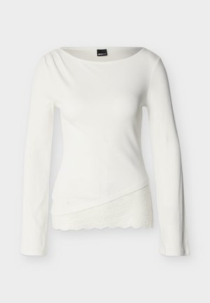 Hvit, langermet, tettsittende topp med asymmetrisk kant og blonder, samt en lett drapert halsringning, laget av ribbestrikket stoff.