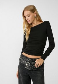 Black long-sleeve top with a scoop neckline, textured fabric, and gathering at the side, paired with dark denim jeans and a studded black belt.