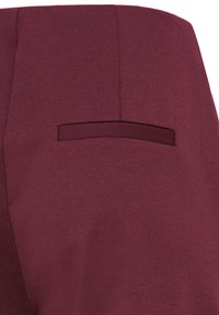 Gros plan d'une poche arrière sur un pantalon sur mesure bordeaux avec un détail de poche passepoilée simple et une texture fine du tissu.