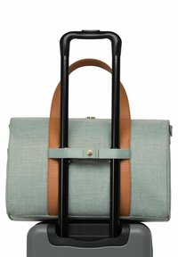 Sac en tissu bleu clair avec des accents en cuir marron, fixé sur la poignée d'une valise noire avec un détail en bouton en métal. Surface texturée et forme structurée.