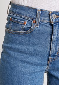 Jean en denim bleu à taille haute, avec des accessoires en cuivre, des coutures orange contrastantes et un petit détail de poche avant.