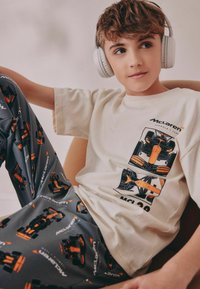 McLaren-tema loungewear sæt bestående af en cremet bomulds T-shirt og grå pyjamasbukser med mønstrede bilgrafikker i orange og sort.