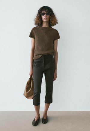Femme aux cheveux bouclés portant des lunettes de soleil, un haut en maille marron à manches courtes, un pantalon noir court, des chaussures noires, tenant un sac marron, debout contre un mur uni.