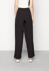 Kvinna sedd bakifrån, klädd i svarta vida byxor, vit kort topp och silverfärgade sneakers mot en enfärgad bakgrund.