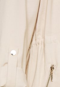 Hellbeige Jacke mit Reißverschluss, silberner Druckknopfschlaufe, elastischem Bund und seitlicher Tasche mit goldener Reißverschlussdetails.