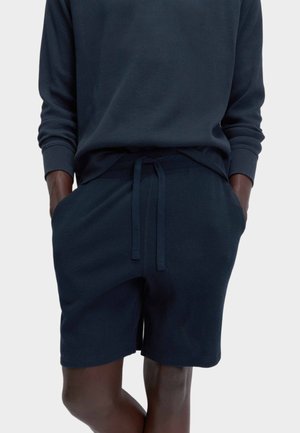 Person trägt ein dunkelblaues Langarmshirt und passende Shorts mit Kordelzug, die Hände in den Taschen, steht vor einem schlichten hellen Hintergrund.