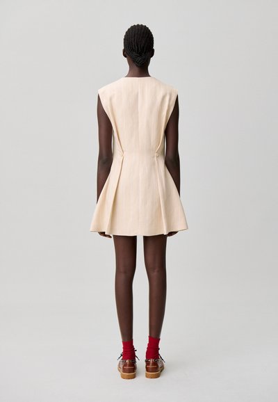 Vestido sin mangas beige con rayas verticales, falda acampanada y dos botones en la cintura. La modelo lleva calcetines rojos y zapatos marrones.