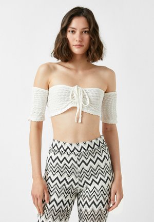 Hvid cropped top med off-the-shoulder design, lavet af tekstureret stof og med en front knude detalje, parret med bukser i sort-hvid zigzag mønster.