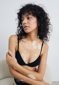 Juodas trikampis braletė su plonomis petnešėlėmis, pagaminta iš �švelnios medžiagos ir su logotipo juostele. Oda rodo šviesius plotus ant pečių ir rankų.