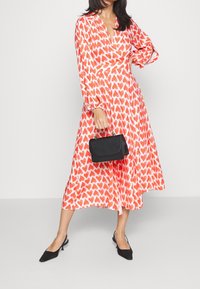 Robe à motifs de cœurs rouges, à manches longues, décolleté en V et jupe fluide. Accessoirisée avec un petit sac à main noir et des escarpins noirs à bout pointu.