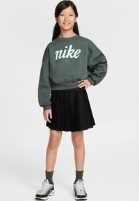 Sweatshirt vert avec logo Nike blanc, jupe plissée noire, chaussettes grises et baskets argentées. Le mannequin se tient avec la main gauche sur la hanche.
