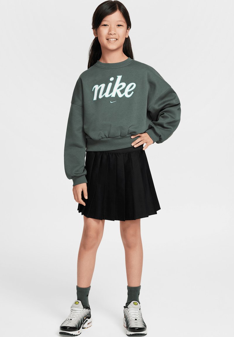 Sweatshirt vert avec logo Nike blanc, jupe plissée noire, chaussettes grises et baskets argentées. Le mannequin se tient avec la main gauche sur la hanche.