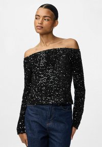 Femme portant un haut noir à épaules dénudées à manches longues recouvert de sequins, avec un jean bleu, regardant vers sa gauche devant un fond uni.