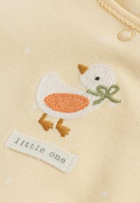 Tissu jaune à pois blancs avec un canard brodé au ventre orange, bec marron et un nœud vert, au-dessus d'une étiquette portant l'inscription « little one ».