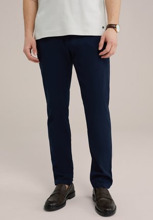 Dunkelblaue Chinos mit strukturierter Oberfläche, kombiniert mit dunkelbraunen Slippern. Das Outfit wird mit einem hellen Hemd abgerundet.